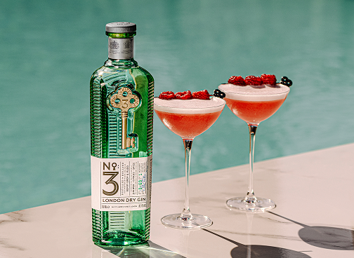 A bottle of No. 3 London Dry Gin placed beside two pink gin cocktails garnished with raspberries and blackberries, set on a marble surface by a pool.